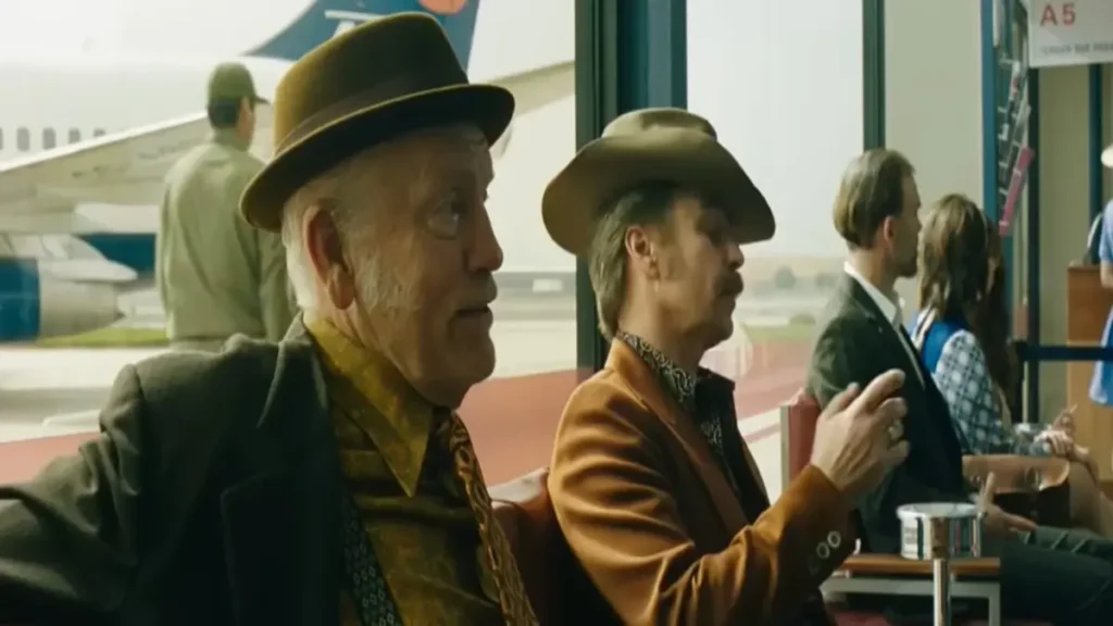 Two men in vintage western attire sitting in an airport terminal in Wild Horse Nine official trailer scene